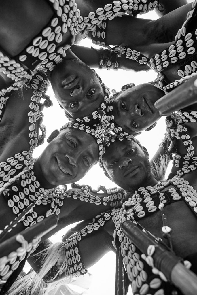 Group of African men smiling in traditional attire adorned with cowrie shells, captured from low angle.